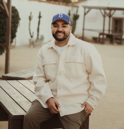 man sitting on a table with a white shirt, brown pants, and blue hat