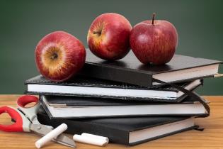 Four black books stacked with 3 apples on top of them placed on a table in front of a chalkboard.